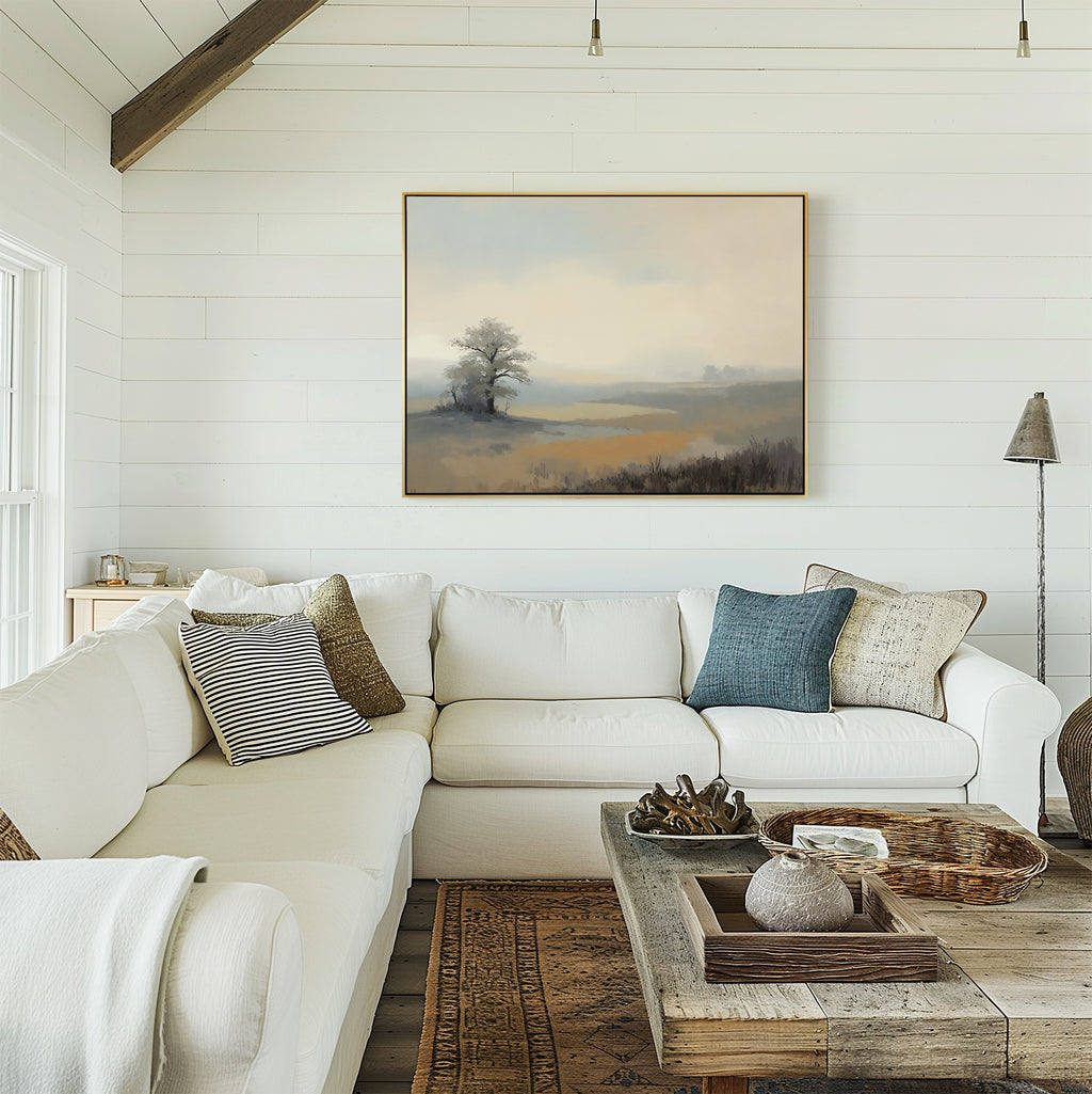 A cozy living room with a white sectional sofa, neutral and blue throw pillows, a rustic wooden coffee table with decor, and the Foggy Solitude Canvas Art - Horizontal featuring a misty landscape of tree and fields on a white shiplap wall.
