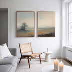A modern living room with a light gray sofa, wooden chair, round white table, and three candles. "Foggy Solitude - Set of 2" art pieces add tranquil decor with misty landscapes and trees on a white wall by large windows.