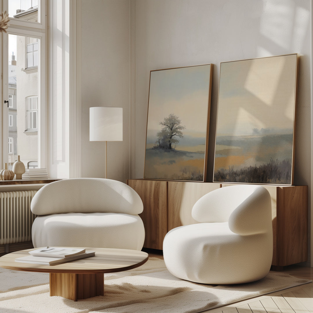 A modern living room with two white chairs, a wooden coffee table, and serene "Foggy Solitude - Set of 2" canvas prints displayed on a sideboard. Sunlight filters in through tall windows, illuminating the tranquil space.
