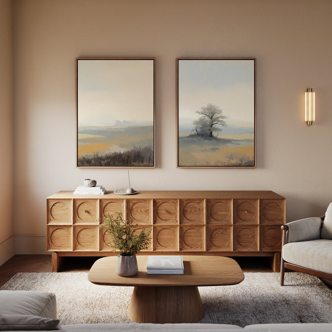 A modern living room with a wooden coffee table, matching sideboard, light sofa, and Foggy Solitude - Set of 2 misty landscape art above the sideboard. A small plant and books decorate the table and sideboard.