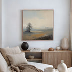 A serene living room with a neutral palette features a soft beige sofa, wooden sideboard with vases and books, and the Foggy Solitude Canvas Art - Square—showing a tree and fields—adding tranquility to the space.