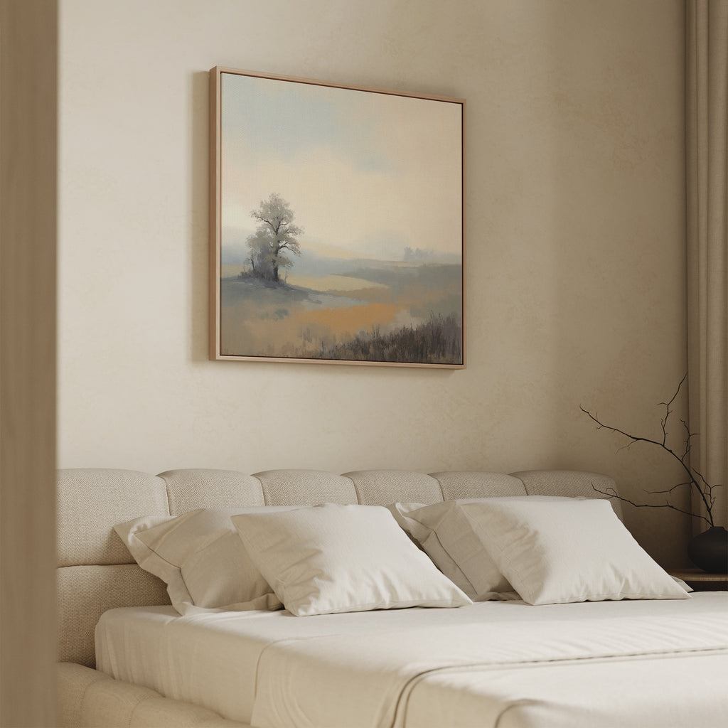 A cozy bedroom features a beige upholstered bed with white pillows and a soft blanket. Above the bed hangs the Foggy Solitude Canvas Art - Square, showcasing a tranquil foggy landscape with a lone tree and misty hills.