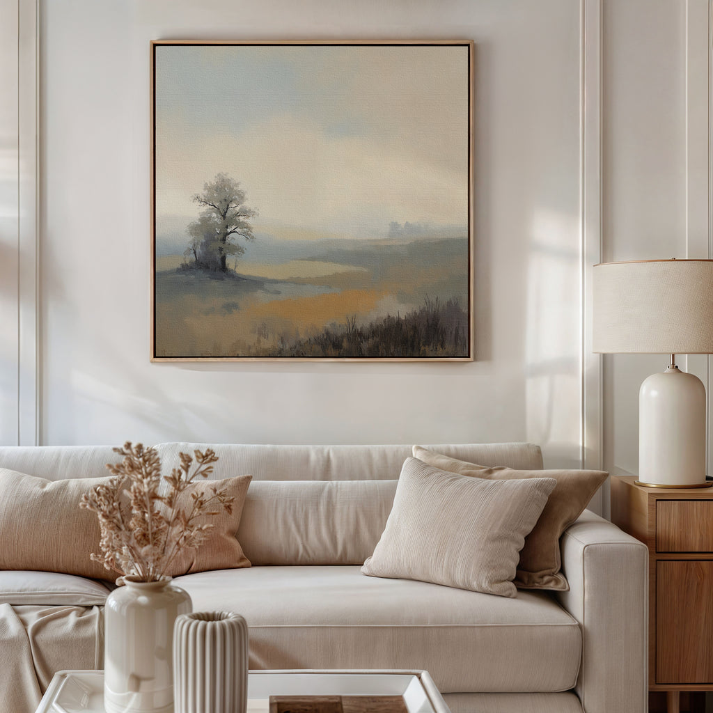 A modern living room with a beige sofa, neutral cushions, a side table with a lamp, and the Foggy Solitude Canvas Art - Square—a tranquil wall art of misty hills and trees—hanging above the sofa.