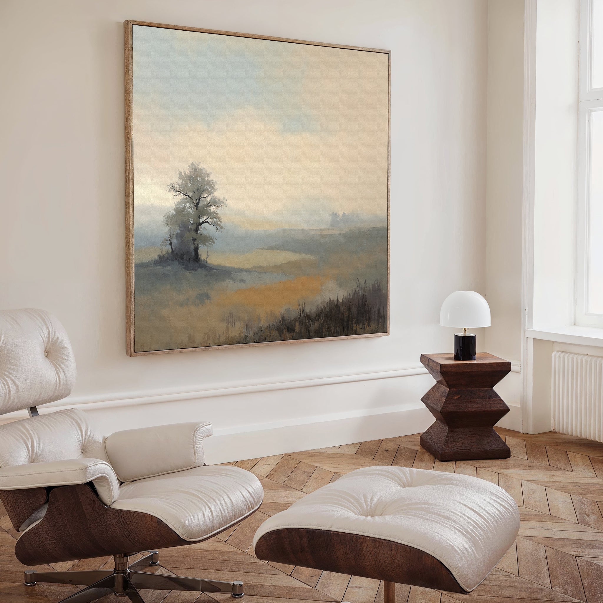 A modern living room with a white leather lounge chair and ottoman, a geometric wooden side table, and the "Foggy Solitude Canvas Art - Square" tranquil wall art of a misty landscape hanging above.