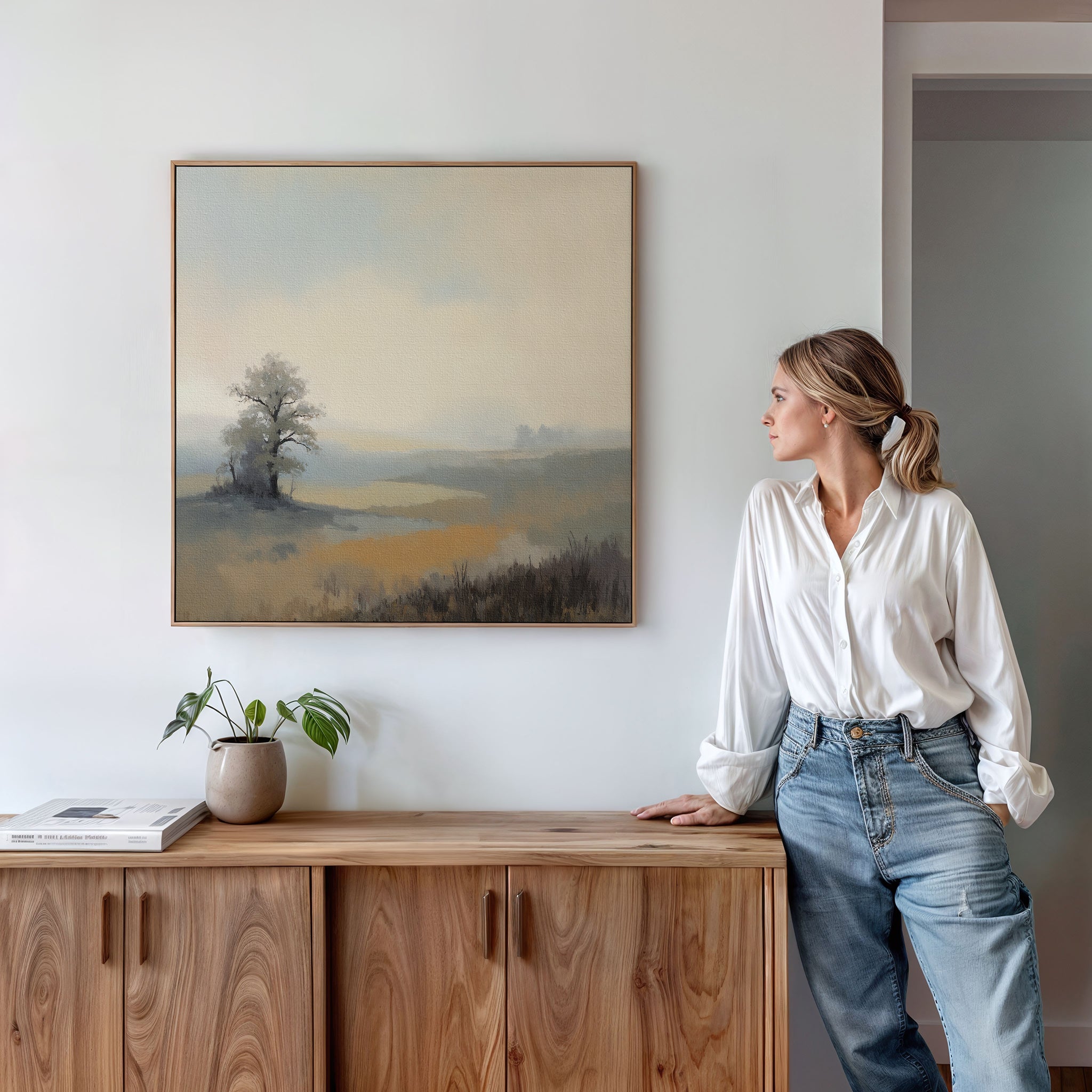 A woman in a white blouse and jeans stands by a wooden sideboard, admiring Foggy Solitude Canvas Art - Square. This tranquil wall art brings calm to the space, complemented by books and a potted plant for a Nordic Park-inspired look.