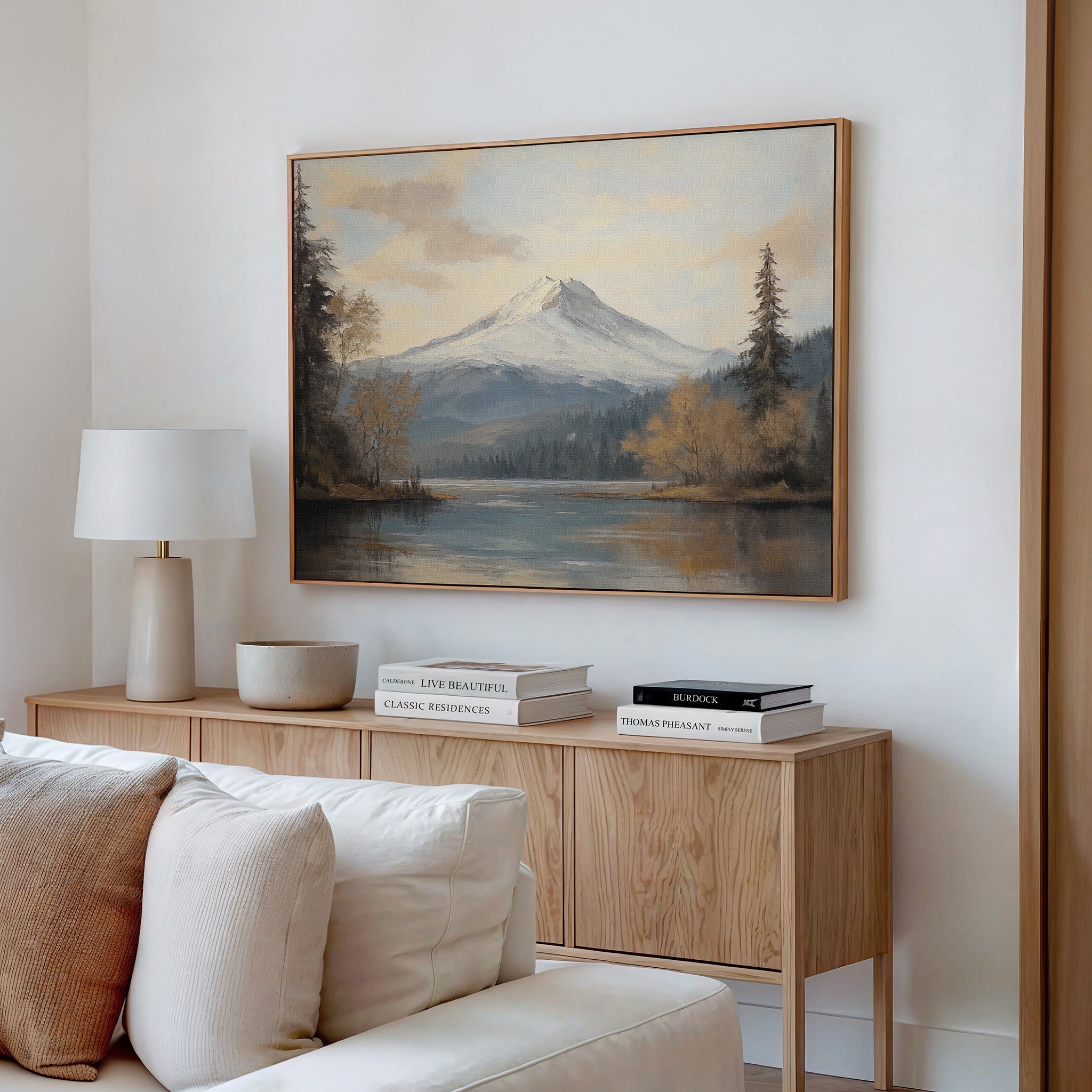 A cozy living room features a light wood sideboard topped with decorative books and a white lamp, while the Frosted Ascent Canvas Art - Horizontal hangs above, capturing tranquil natural beauty. A cream sofa is partially visible in front.