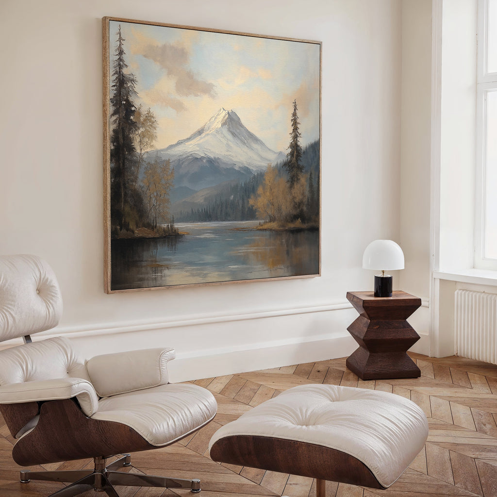 A modern living room displays the Frosted Ascent Canvas Art - Square above a white lounge chair and ottoman. The serene artwork of snowy mountains, trees, and a lake is illuminated by light streaming through tall windows.