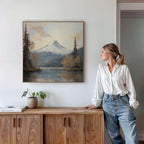 A woman in a white blouse and jeans stands by a wooden cabinet, admiring the Frosted Ascent Canvas Art - Square, a serene painting of a snow-capped mountain and lake, with a plant and books resting on the cabinet.