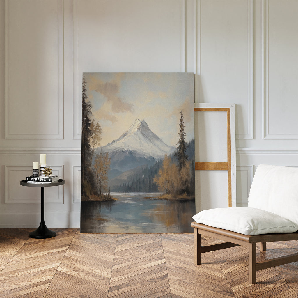 Frosted Ascent Canvas Art - Vertical, featuring a snow-capped mountain by a lake, rests against a white-paneled wall in a bright room with herringbone wood floors, a cushioned chair, candles on a small table, and an empty frame.