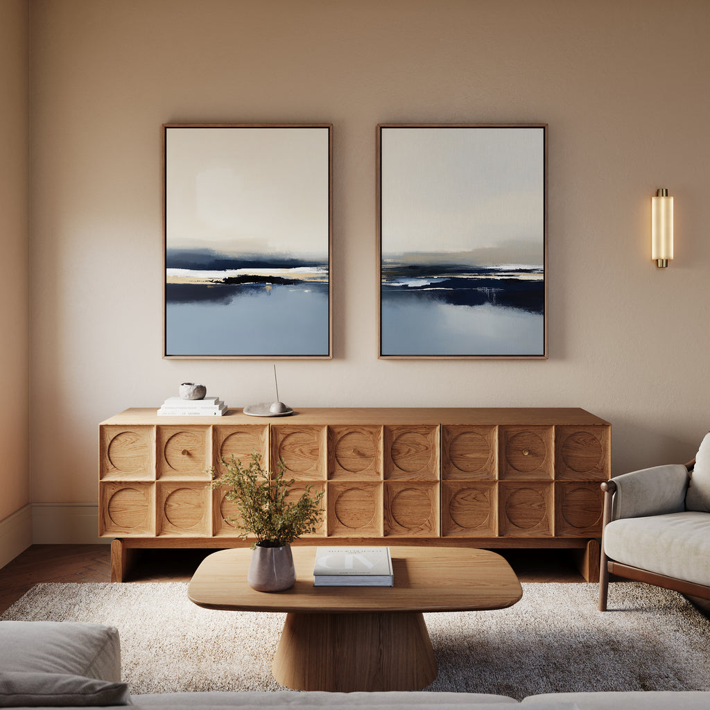 A modern living room features a wooden coffee table, matching sideboard, light sofa, and the Frozen Bay - Set Of 2 artworks above. A vase with greenery sits on the table; soft lighting enhances the cozy modern decor.