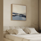 A minimalist bedroom features a beige tufted bed with four white pillows, a soft blanket, and the Frozen Bay Canvas Art - Square hanging above. A bare branch in a vase decorates the nightstand.