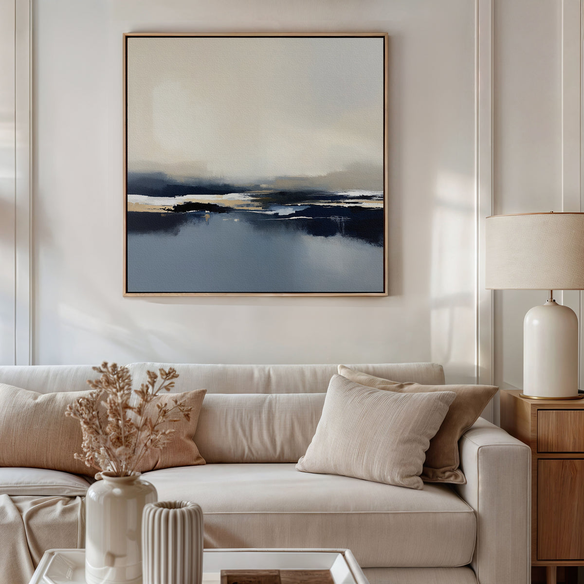 A modern living room with a beige sofa, neutral cushions, a side table with a ceramic lamp, dried flowers in a vase, and the Frozen Bay Canvas Art - Square featuring blue and cream tones on the wall.