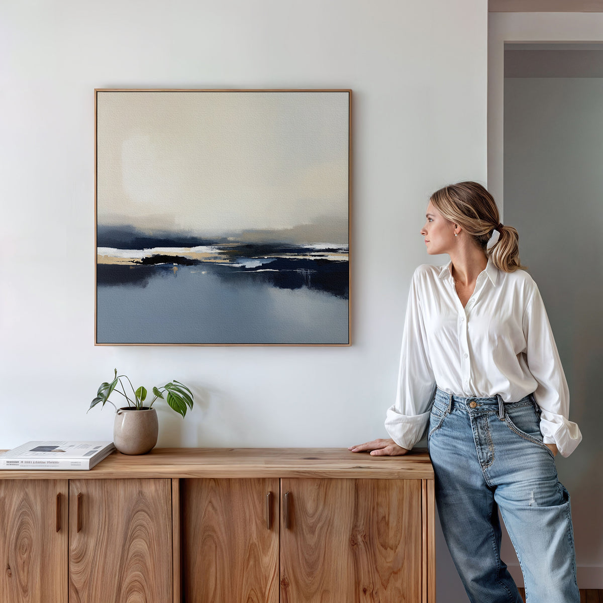 A woman in a white blouse and jeans leans on a wooden cabinet, gazing at the Frozen Bay Canvas Art - Square above her—an ideal modern living room accent. A small potted plant and a book sit on the cabinet.