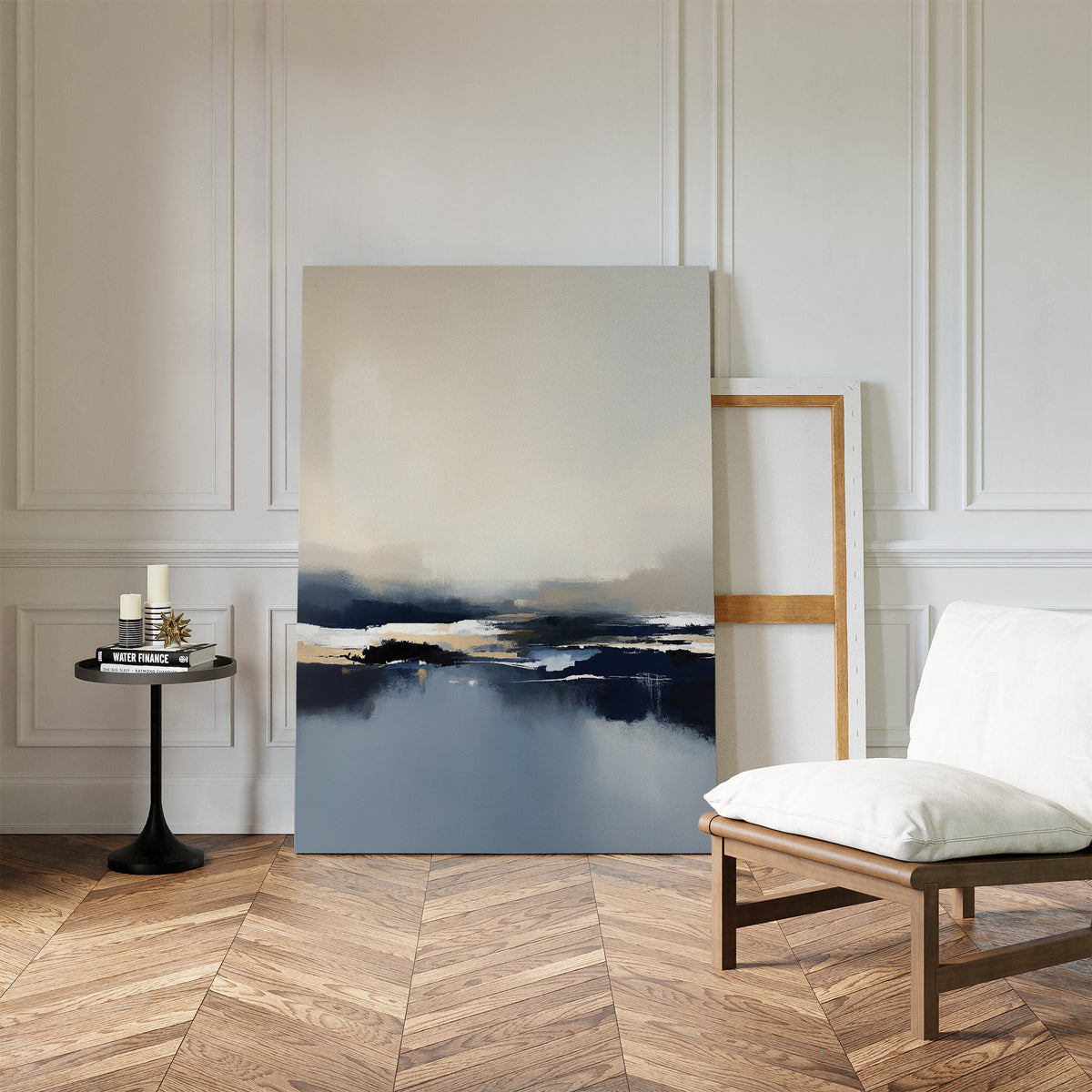 A minimalist room with herringbone wood floors, a white cushioned chair, a round black side table with candles, and the Frozen Bay Canvas Art - Vertical—showcasing blue, white, and beige tones—leaning against a white paneled wall.