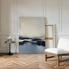 A minimalist room with herringbone wood floors, a white cushioned chair, a round black side table with candles, and the Frozen Bay Canvas Art - Vertical—showcasing blue, white, and beige tones—leaning against a white paneled wall.