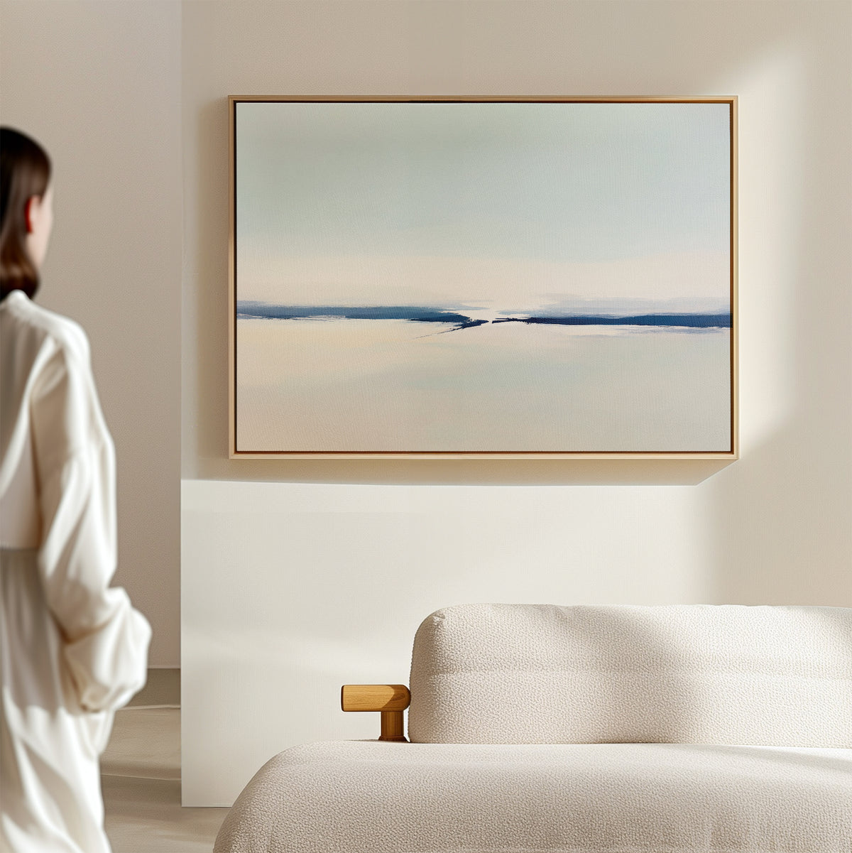 A person stands in a bright, minimal room, admiring the "Frozen Channel Canvas Art - Horizontal" in soft blues and creams hung above a beige sofa—an ideal addition to modern living room decor.