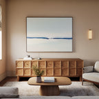 A minimalist living room with a light wood coffee table, matching sideboard, beige sofa, neutral rug, wall-mounted light, and the Frozen Channel Canvas Art - Horizontal in soft blue and white tones above the sideboard for modern decor.