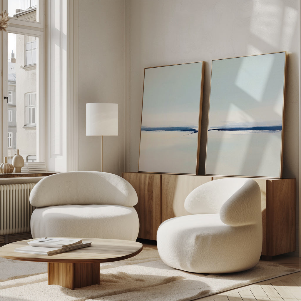 A modern living room with the Frozen Channel - Set Of 2 white curved armchairs, a low wooden coffee table, and serene blue and beige art prints resting on a wooden sideboard by a sunlit window.