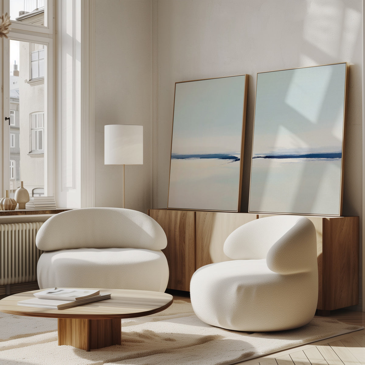 A modern living room with the Frozen Channel - Set Of 2 white curved armchairs, a low wooden coffee table, and serene blue and beige art prints resting on a wooden sideboard by a sunlit window.