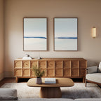 A serene modern living room features the Frozen Channel - Set Of 2 wall art above a wooden sideboard, a low coffee table with books and a plant, a neutral armchair, and sconce lighting for warmth.