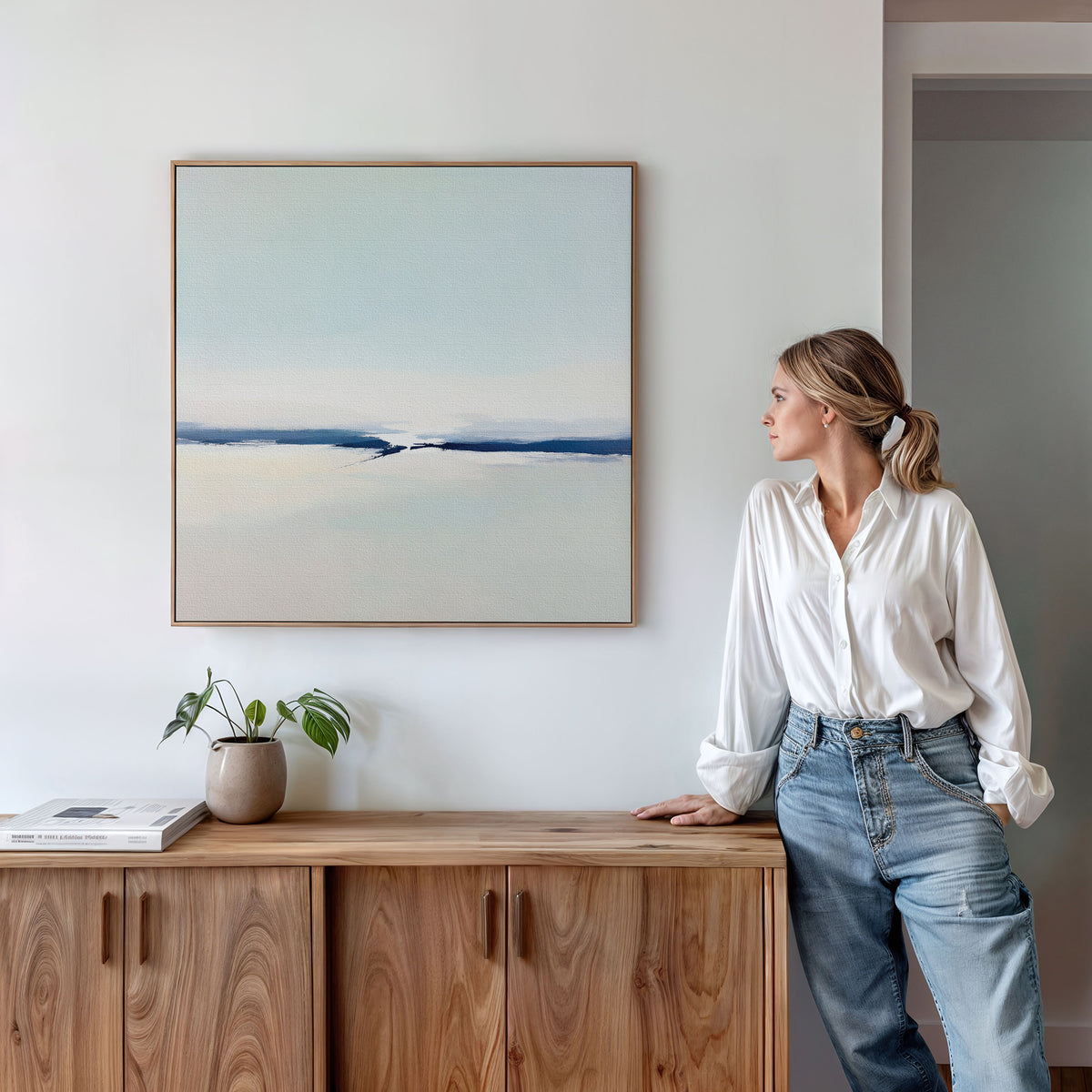 A woman in a white blouse and jeans leans against a wooden cabinet, admiring the Frozen Channel Canvas Art - Square on the wall. A small potted plant and books complete this stylish modern living room setup.