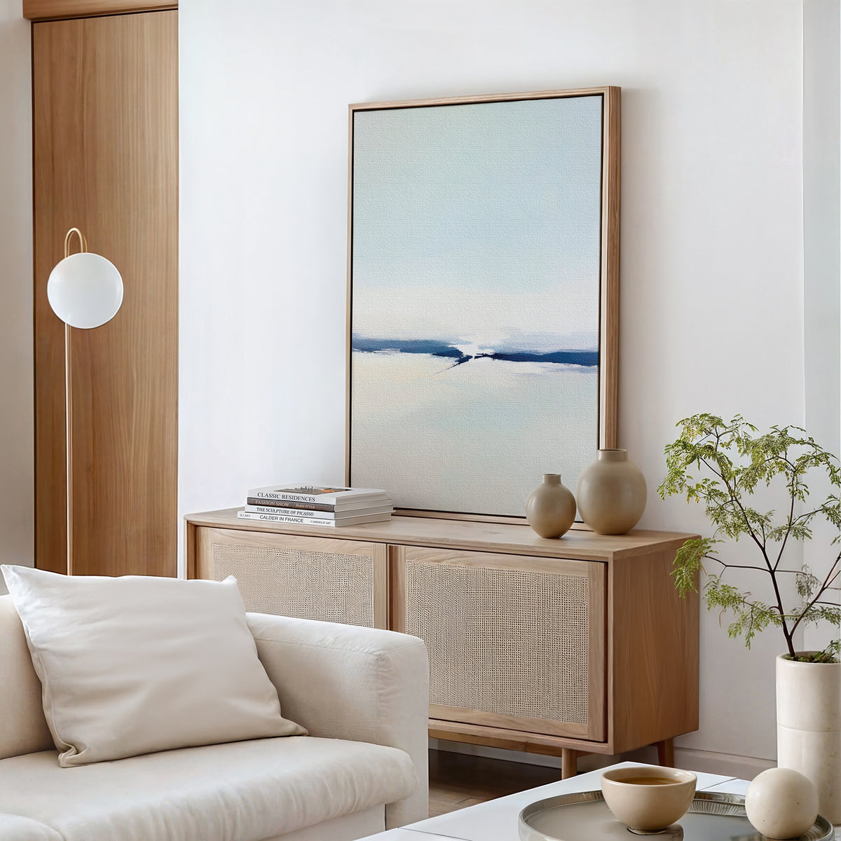 A minimalist living room with modern decor features the Frozen Channel Canvas Art - Vertical, a light wood sideboard, ceramic vases, stacked books, a white sofa with pillows, a round hanging light, and a potted plant for a calming atmosphere.