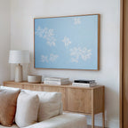 A light wood sideboard with stacked books, a lamp, and a bowl sits against a white wall beneath the Gentle Blossoms Canvas Art - Horizontal. In front, there’s a beige couch with cushions. The space features minimalist, modern decor.