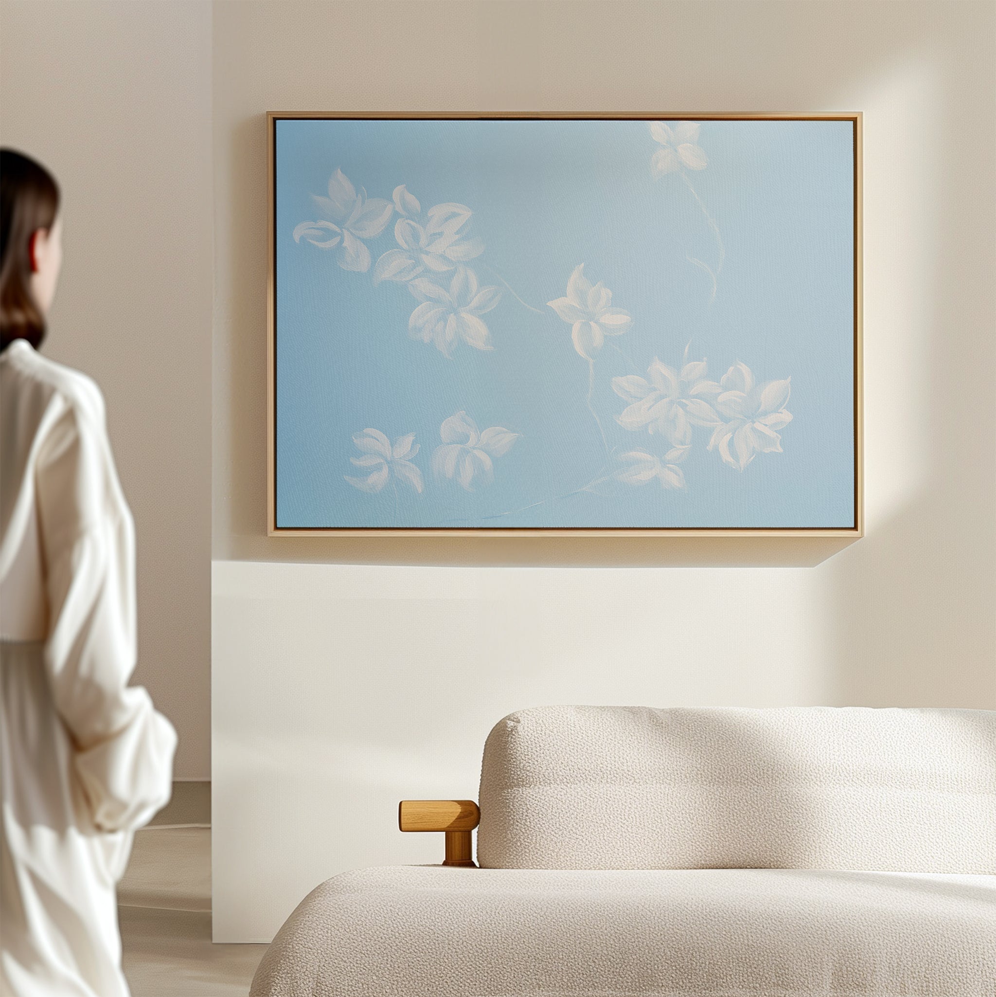 A person in white stands in a minimal room, gazing at the Gentle Blossoms Canvas Art - Horizontal, featuring white floral motifs on a light blue background above a beige textured sofa, softly lit for a serene ambiance.