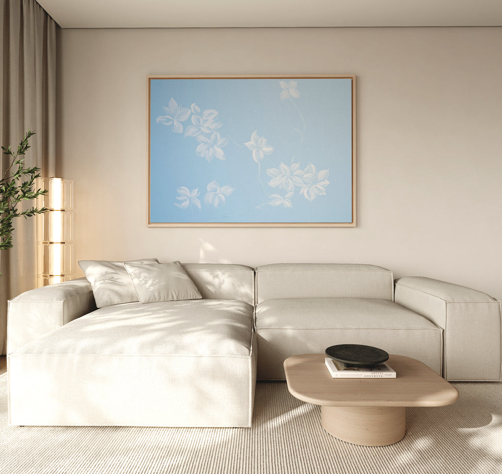 A modern living room with a beige sectional, light wood coffee table, and the Gentle Blossoms Canvas Art - Horizontal featuring blue and white florals. Neutral tones and natural light contribute to a calm atmosphere.