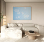A modern living room with a beige sectional, light wood coffee table, and the Gentle Blossoms Canvas Art - Horizontal featuring blue and white florals. Neutral tones and natural light contribute to a calm atmosphere.