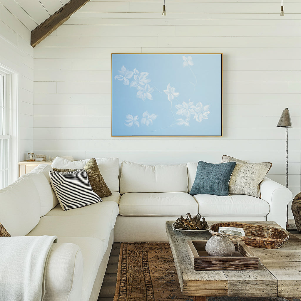 A bright, airy living room with white shiplap walls, a large white sectional sofa with assorted pillows, a rustic wooden coffee table, and the Gentle Blossoms Canvas Art - Horizontal featuring blue florals above the couch.