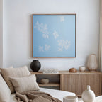A minimalist living room with a beige sofa, neutral cushions, a wooden sideboard with decor, and the Gentle Blossoms Canvas Art - Square, a large light blue canvas featuring white floral designs, displayed on the wall.