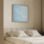 A minimalist bedroom features a beige upholstered bed, white bedding, and the Gentle Blossoms Canvas Art - Square above the headboard. A leafless branch in a vase accents the bedside table.