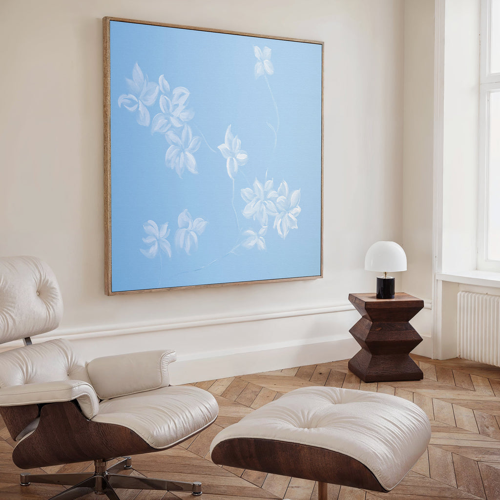 A modern living room featuring a white leather lounge chair and ottoman, a dark wood side table with a white lamp, and the Gentle Blossoms Canvas Art - Square as striking floral wall art. Light wooden floors and a large window complete the look.