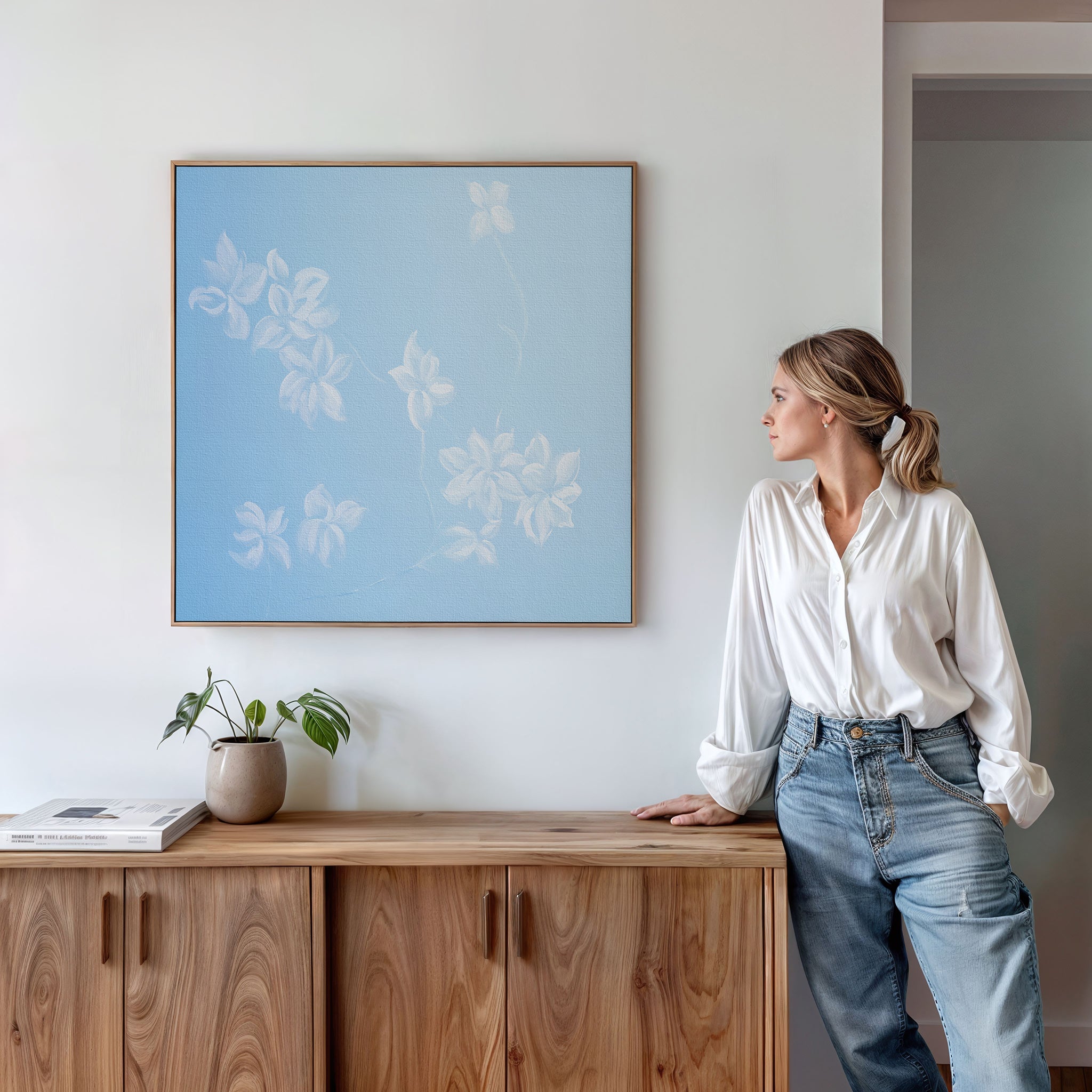 A woman in a white blouse and jeans leans against a wooden cabinet, looking at the Gentle Blossoms Canvas Art - Square hanging on the white wall above her. A plant and books decorate the cabinet.