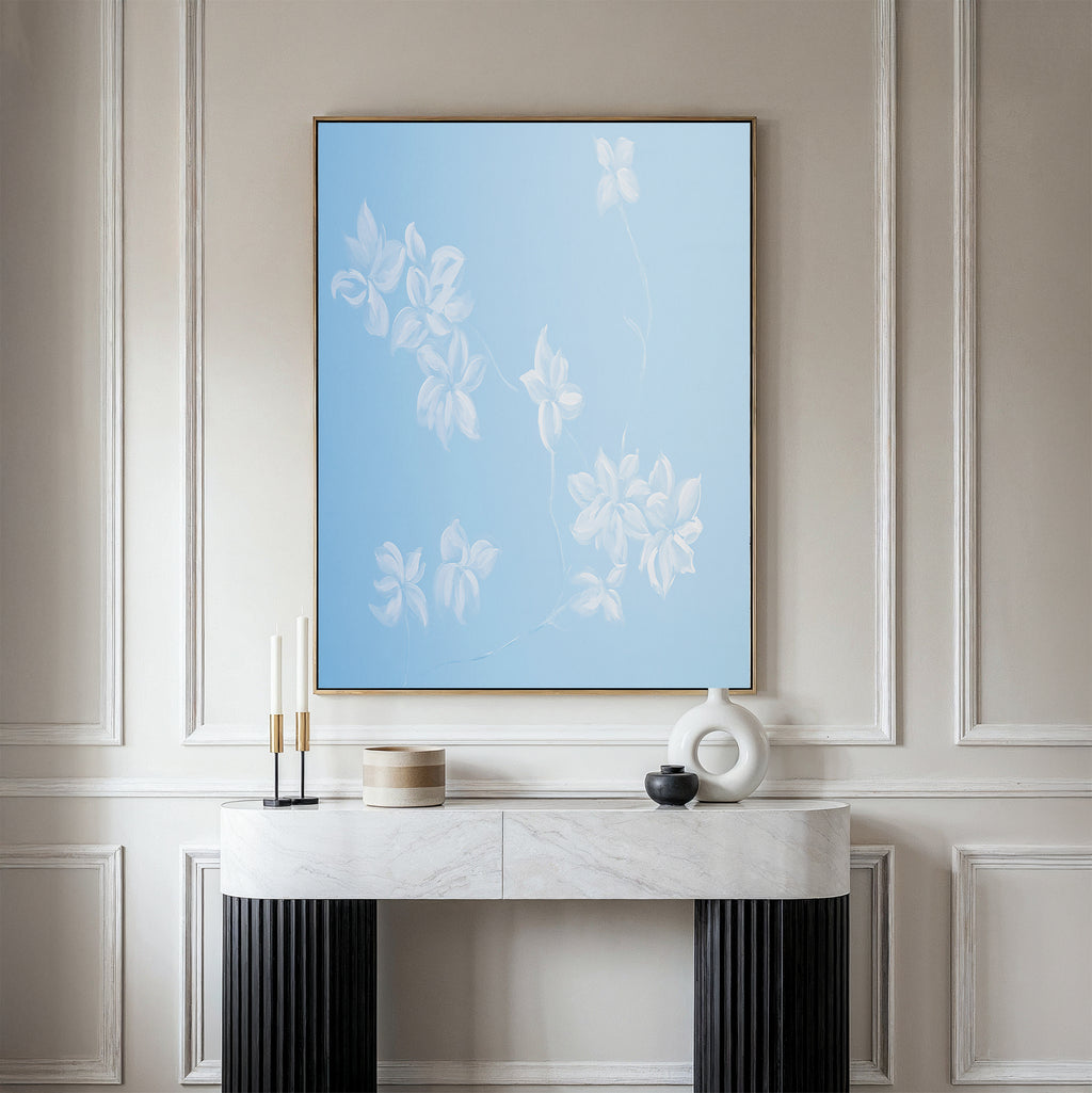 A minimalist console table with decor stands before a beige paneled wall, highlighted by the Gentle Blossoms Canvas Art - Vertical, featuring delicate white flowers in powder blue tones.