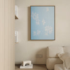 A minimalist living room features a beige sofa, a light wood shelf with books, a geometric white decor piece, and the Gentle Blossoms Canvas Art - Vertical in powder blue with white outlines on a cream-colored wall.
