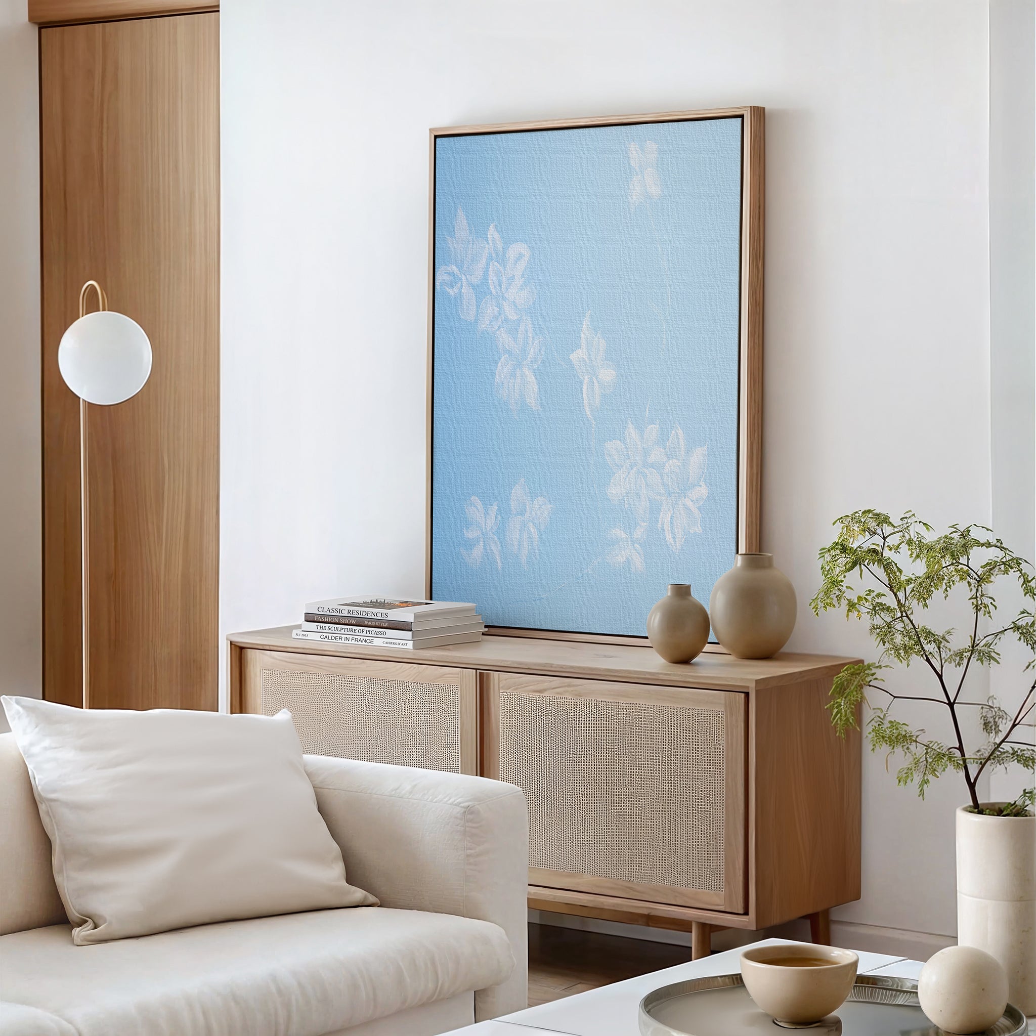 A modern living room with a light wood sideboard, the Gentle Blossoms Canvas Art - Vertical, stacked books, vases, a round lamp, a white sofa with a pillow, a coffee table, and a small green potted plant.
