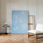 The Gentle Blossoms Canvas Art - Vertical, featuring powder blue with white floral designs, leans against a white paneled wall beside an empty wooden frame. Nearby, a cushioned chair and small table with books rest on the chevron wood floor.