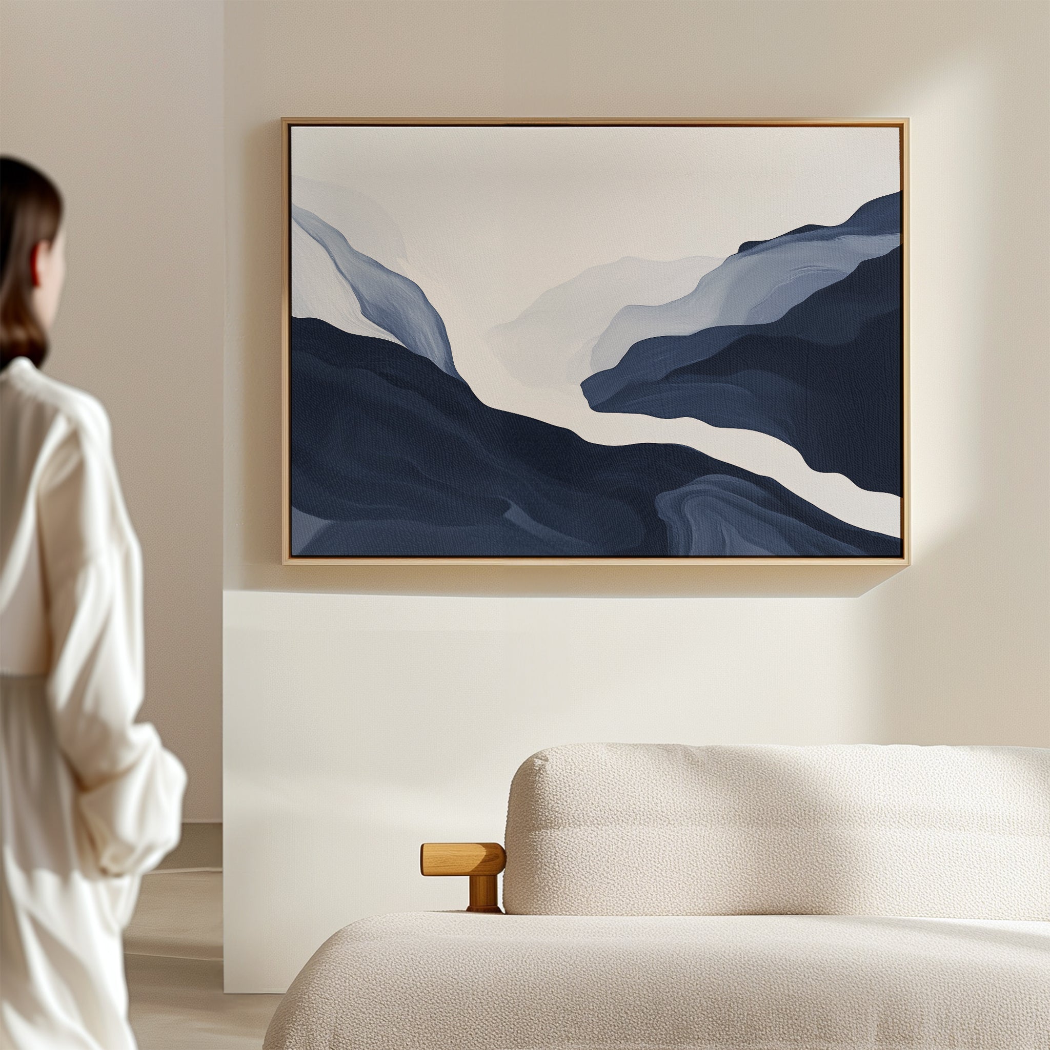 A woman in a white shirt stands in a minimalist bedroom, admiring the Glacier Flow Canvas Art - Horizontal with navy blue wavy shapes above a beige textured bed—an elegant touch of modern home decor.