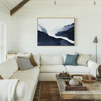 A bright, cozy living room with modern decor: a white sectional sofa, assorted pillows, a rustic wooden coffee table, and the Glacier Flow Canvas Art - Horizontal in blue and white on a shiplap wall. Natural light streams in from the left.