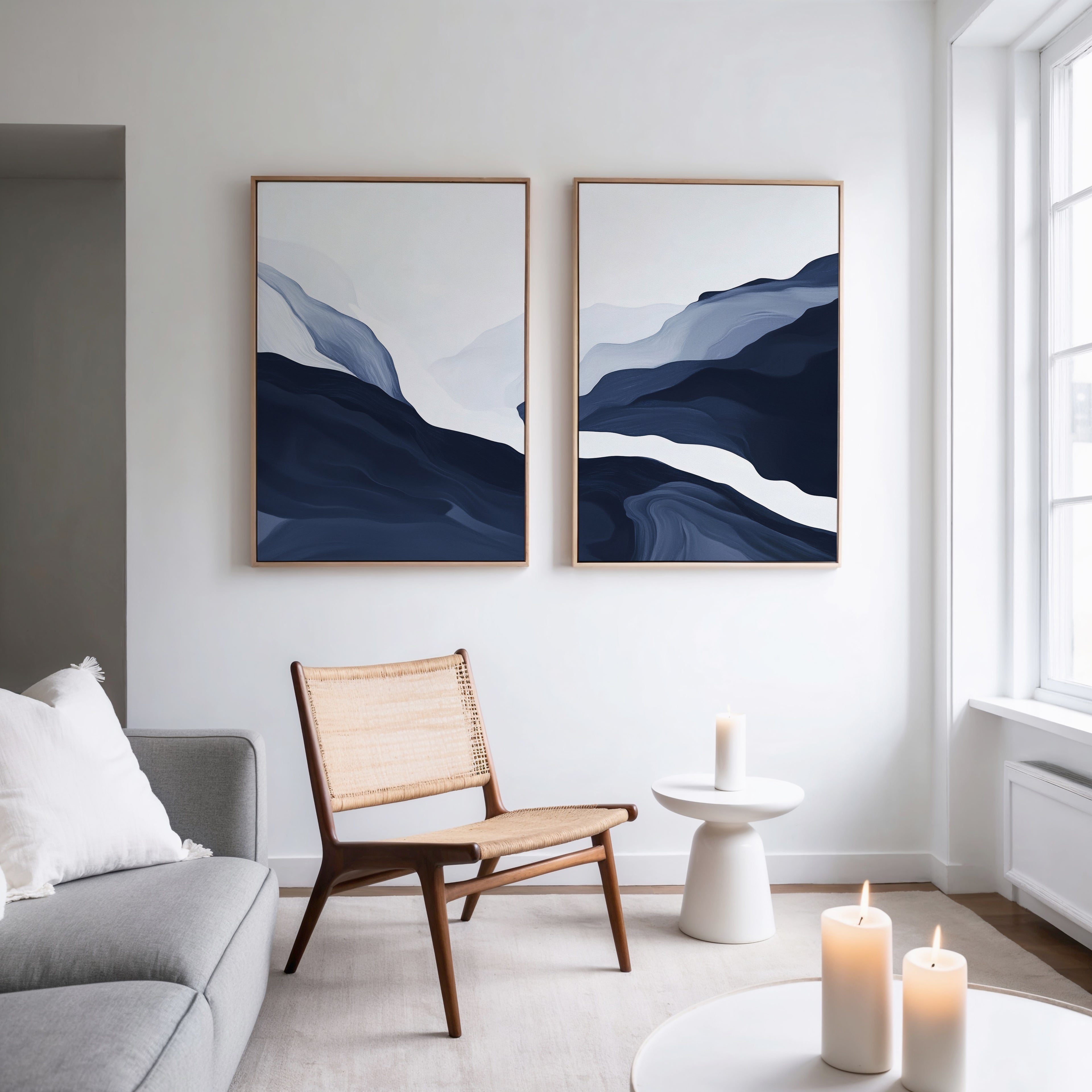 A modern, minimalist living room with a light gray sofa, wooden chair, small round side table, candles, and Nordic decor featuring the Glacier Flow - Set Of 2 abstract wall art in blue and white tones on a white wall near a large window.