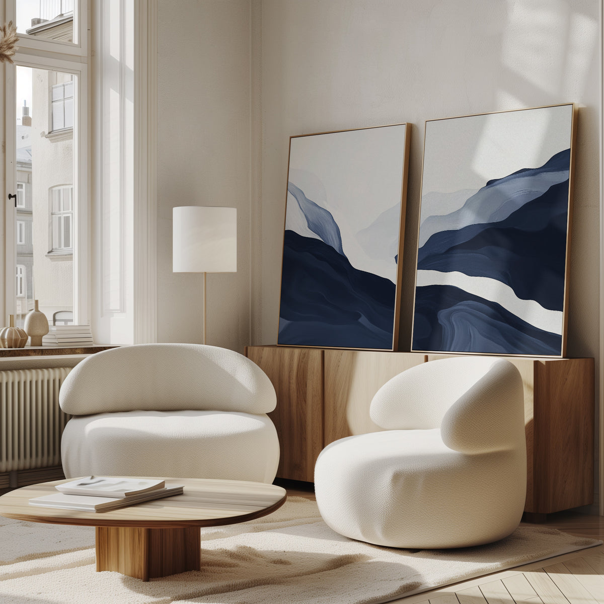 A modern living room with Nordic decor features the Glacier Flow - Set Of 2 rounded white chairs, a low wooden coffee table, a wooden sideboard, abstract navy blue wall art leaning against the wall, and large windows that bring in ample natural light.