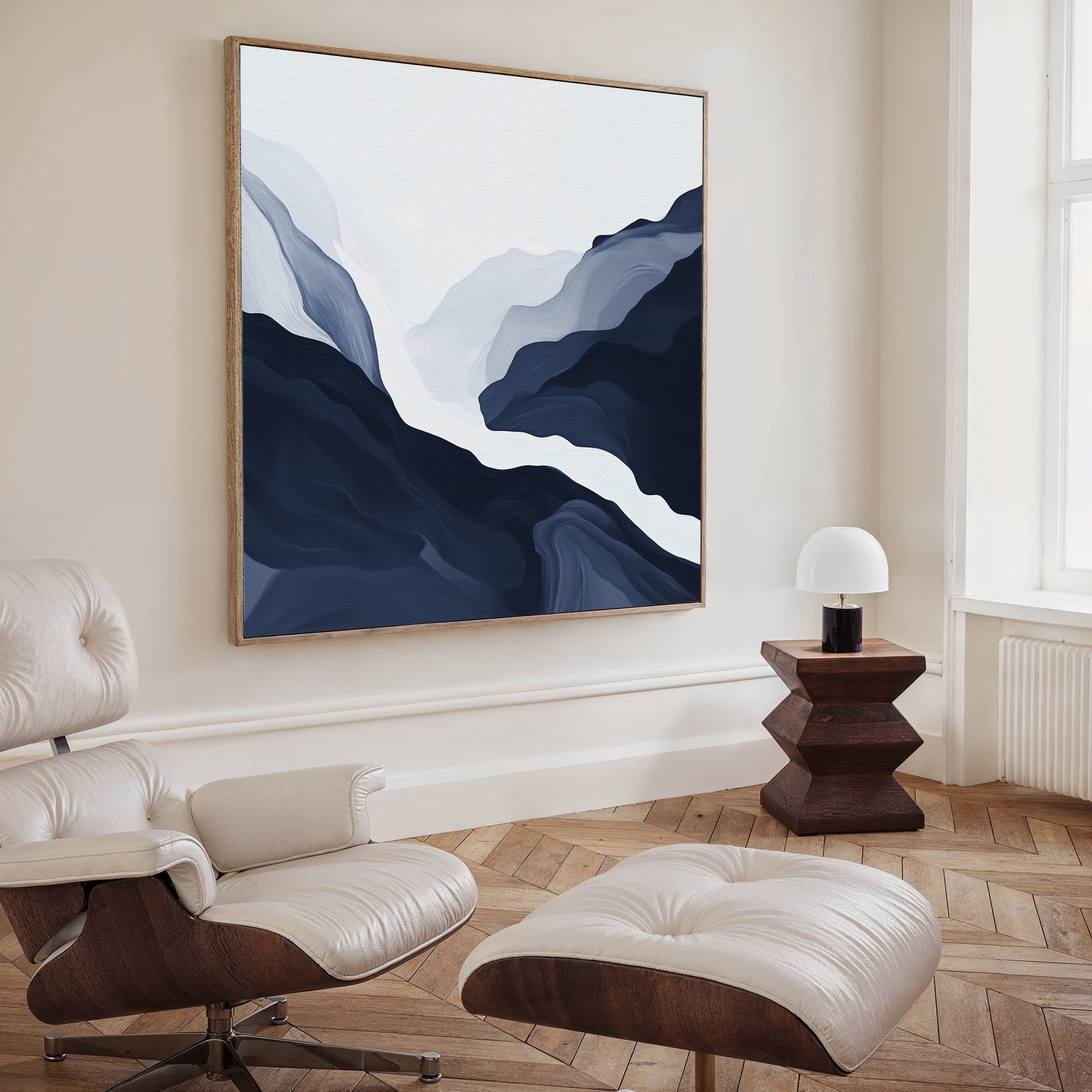 A modern living room features a white lounge chair with ottoman, a wooden side table with a white lamp, and the Glacier Flow Canvas Art - Square depicting abstract mountains on the navy wall. Sunlight streams in through a tall window, enhancing the decor.