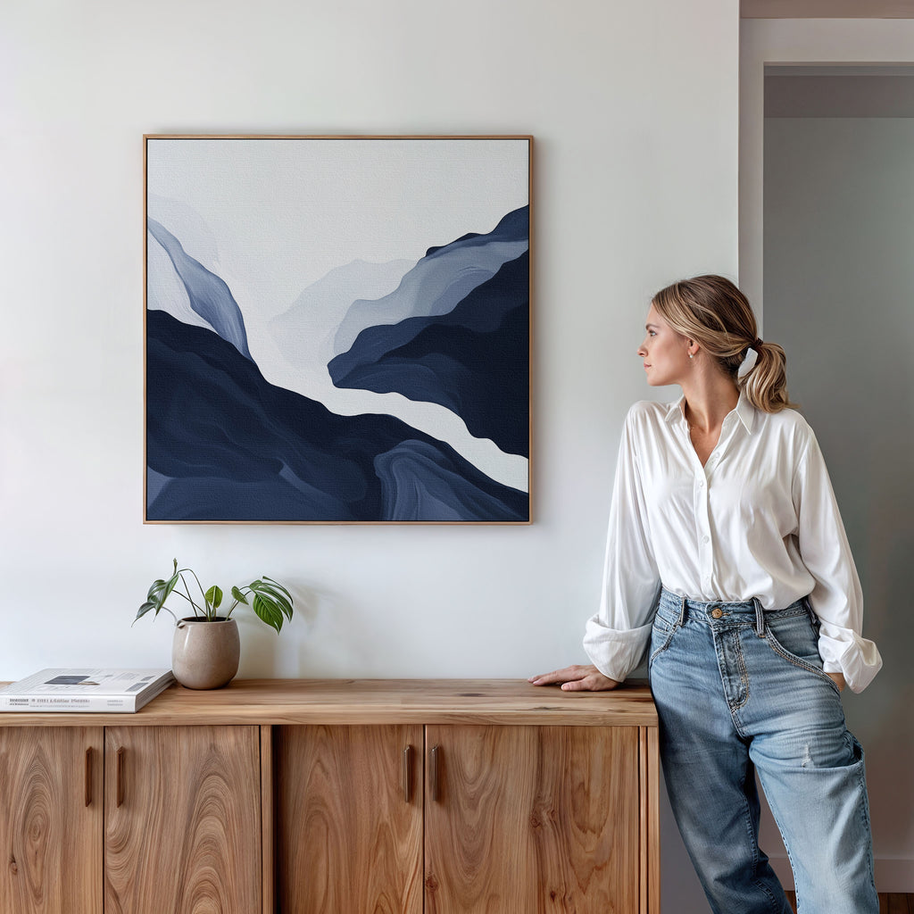 A woman in a white shirt and jeans stands by a wooden sideboard, admiring the Glacier Flow Canvas Art - Square in blue and white above her. A small potted plant and books add charm to the modern decor.