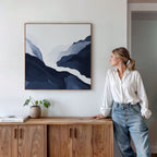 A woman in a white shirt and jeans stands by a wooden sideboard, admiring the Glacier Flow Canvas Art - Square in blue and white above her. A small potted plant and books add charm to the modern decor.