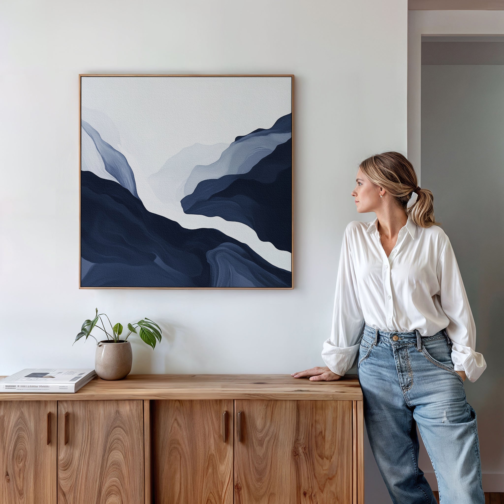 A woman in a white shirt and jeans stands by a wooden sideboard, admiring the Glacier Flow Canvas Art - Square in blue and white above her. A small potted plant and books add charm to the modern decor.