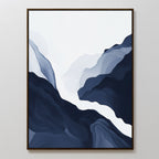 The Glacier Flow Canvas Art - Vertical features abstract flowing shapes in navy blue, gray, and white, evoking a mountain landscape with a river. This modern piece is framed with a thin dark border and displayed on a white wall.