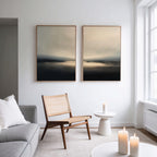 A modern, minimalist living room with a gray sofa, woven chair, “Golden Fog Horizon - Set Of 2” wall art above, a small white side table, and three lit candles for a calm atmosphere.