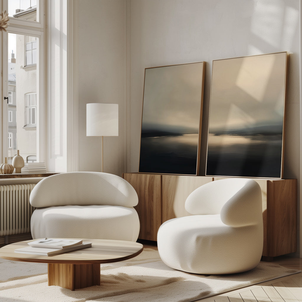 A cozy, modern living room features two white rounded armchairs, a low wooden coffee table, and a sideboard beneath the Golden Fog Horizon - Set Of 2 wall art. Sunlight pours in through tall windows, highlighting the coastal-inspired decor.